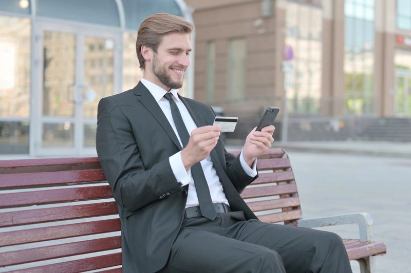 mężczyzna w garniturze siedzący na ławce i trzymający smartfona oraz kartę kredytową