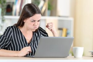 młoda kobieta siedząca przy biurku odgrażająca się pięścią wpatrzona w ekran szarego laptopa