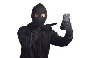 mężczyzna w czarnym ubraniu, rękawiczkach i czarnej kominiarsce wskazujący palcem na telefon trzymany w dłoni