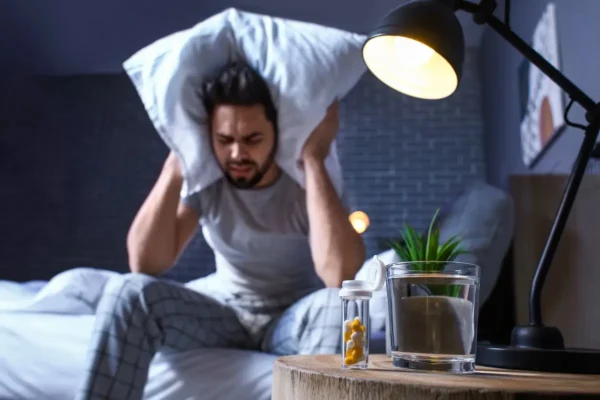 mężczyzna siedzący na łóżku w nocy i zasłaniający uszy poduszką z powodu hałasu