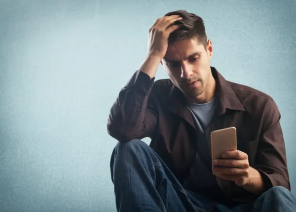 przygnębiony mężczyzna siedzący na podłodze i trzymający telefon z głową opartą na dłoni