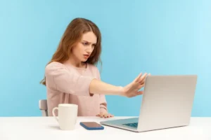 młoda kobieta siedząca na krześle przy stole z szarym laptopem i zasłaniająca się ręką z krzywą miną