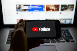 Dłoń trzymająca smartfon z wyświetlonym logo YouTube na tle otwartego laptopa z widocznym panelem filmów, symbolizująca korzystanie z platformy wideo.