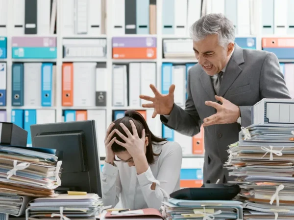 Przełożony krzyczący na pracownicę siedzącą przy biurku, co symbolizuje poniżanie i nękanie w miejscu pracy