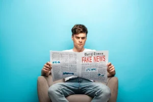 Zmieszany i poirytowany mężczyzna siedzi w fotelu i czyta gazetę z nagłówkiem FAKE NEWS na jasnoniebieskim tle.