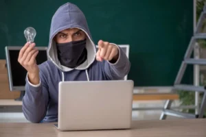 Mężczyzna w bluzie z kapturem i zasłoniętą twarzą siedzi przy laptopie, trzyma żarówkę i wskazuje palcem. Kradzież pomysłu i kodu źródłowego aplikacji.